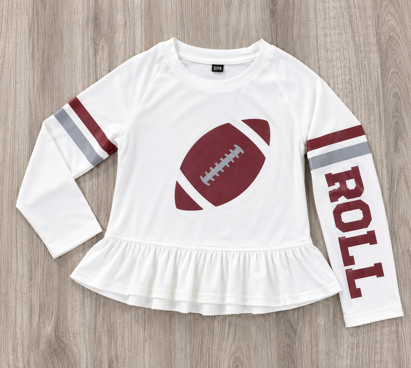 A white long-sleeve t-shirt with a ruffled hem, featuring a crimson football graphic on the chest, crimson and gray stripes on the sleeves, and the word 'ROLL' printed vertically on one sleeve, laid flat on a gray wood background. This is a SPF t-shirt for girls for gameday in Alabama.