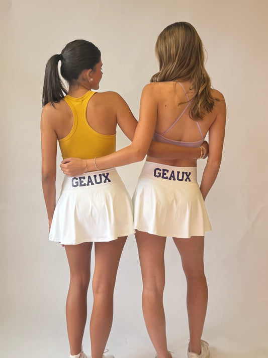 Two women standing side by side from behind wearing matching white game day athletic tennis skirts pleated skirts with 'GEAUX' printed in purple on the back, along with purple and golds sports bras, against a plain light beige studio background.