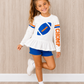 Girls football t-shirt that is spf 50. The girl is wearing the white shirt with a blue football in shimmer color on the front. The shirt has  varsity stripes and says chomp down the sides of them. The girl stand sin front of a wooden wall and on a wooden floor. She is ready for football in Florida. 