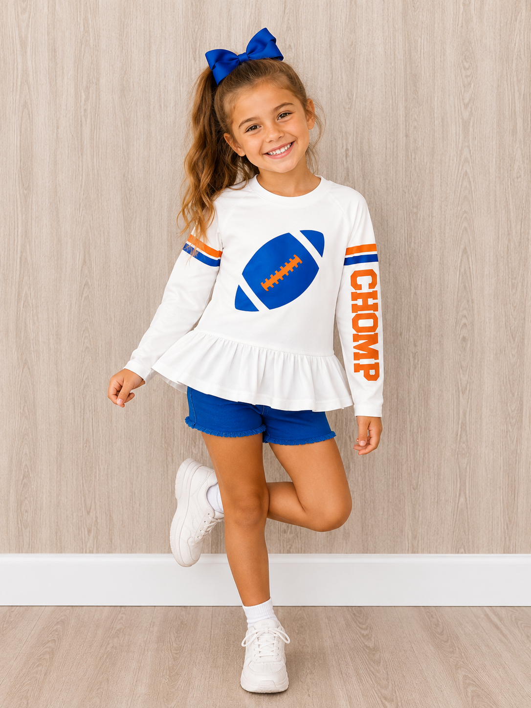 Girls football t-shirt that is spf 50. The girl is wearing the white shirt with a blue football in shimmer color on the front. The shirt has  varsity stripes and says chomp down the sides of them. The girl stand sin front of a wooden wall and on a wooden floor. She is ready for football in Florida. 