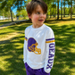 A young boy wearing a white long-sleeve athletic shirt with a purple and gold football helmet graphic and the word 'GEAUX' on the sleeve, paired with purple shorts, standing outdoors on a grassy park lawn. The boy wears a gameday shirt made for Louisiana football. 
