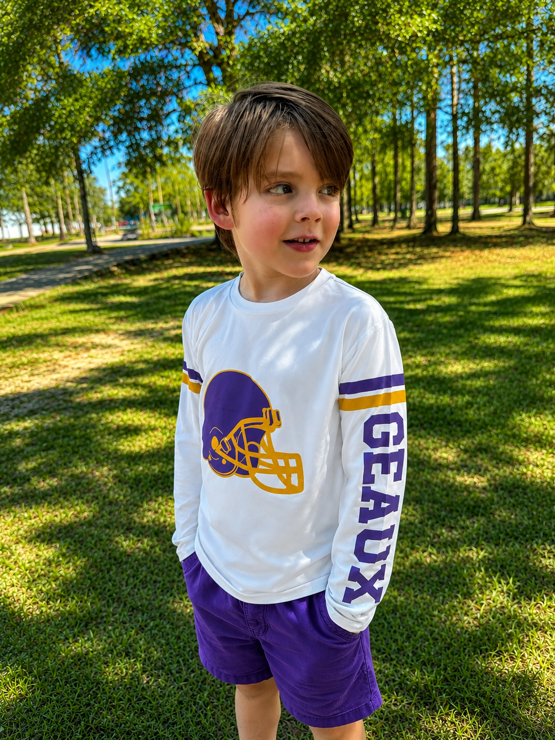 A young boy wearing a white long-sleeve athletic shirt with a purple and gold football helmet graphic and the word 'GEAUX' on the sleeve, paired with purple shorts, standing outdoors on a grassy park lawn. The boy wears a gameday shirt made for Louisiana football. 