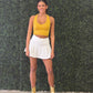 A woman stands in front of a green leafy wall wearing a yellow ribbed crop top, a white tennis skirt, and metallic gold ankle boots. The skirt says geaux on the back which is a popular Louisiana term. The skirt is made for game day. 