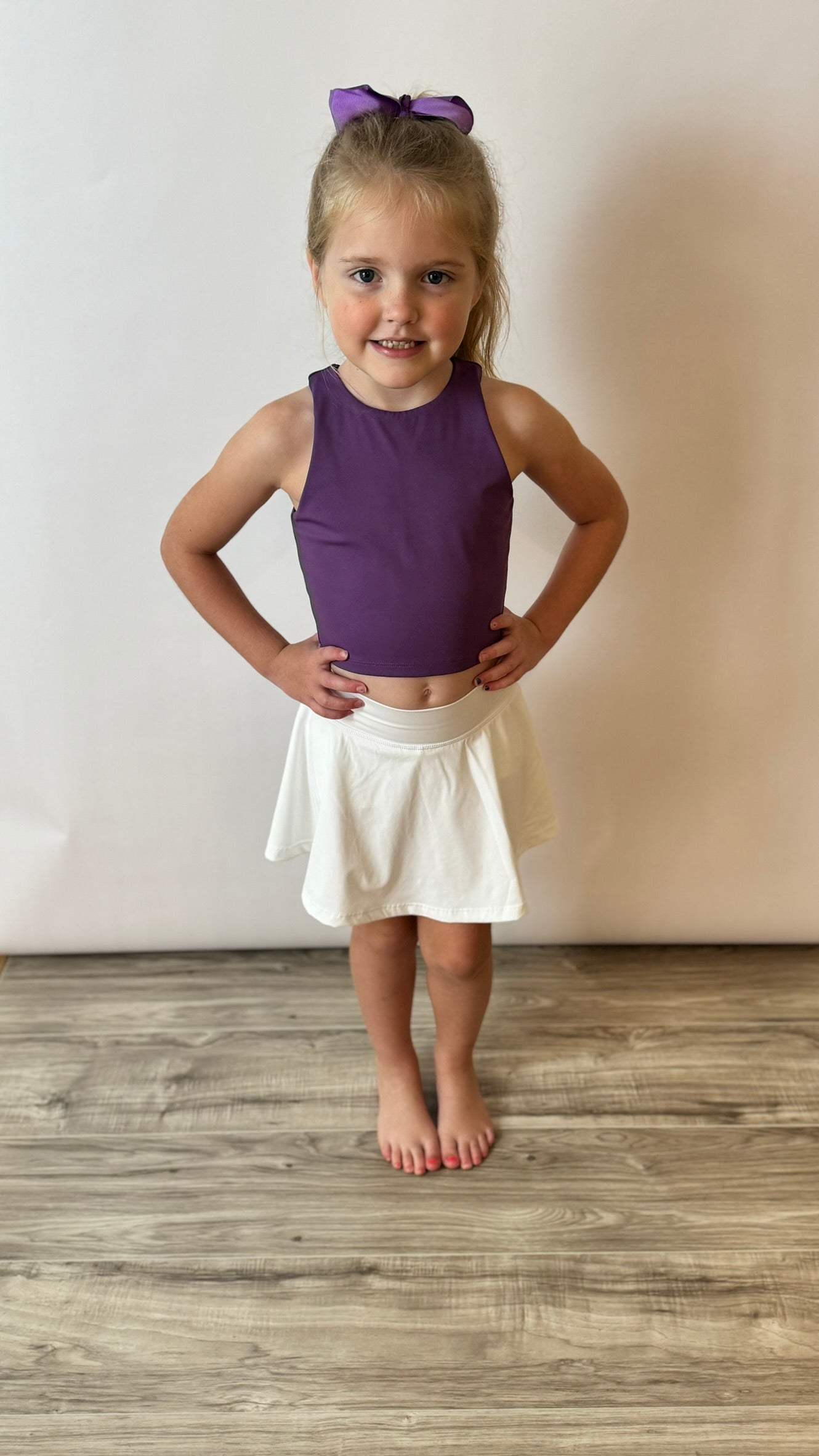 Girl wearing a white athletic skirt with the words geaux on it. She pairs it with a purple athletic top and matching bow. 
