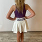 A child wearing a purple and white tennis skirt with 'Geaux' written on the front, and a matching bow. The child has blonde hair and is standing against a white background. This skirt is a game day skirt popular in Louisiana. 