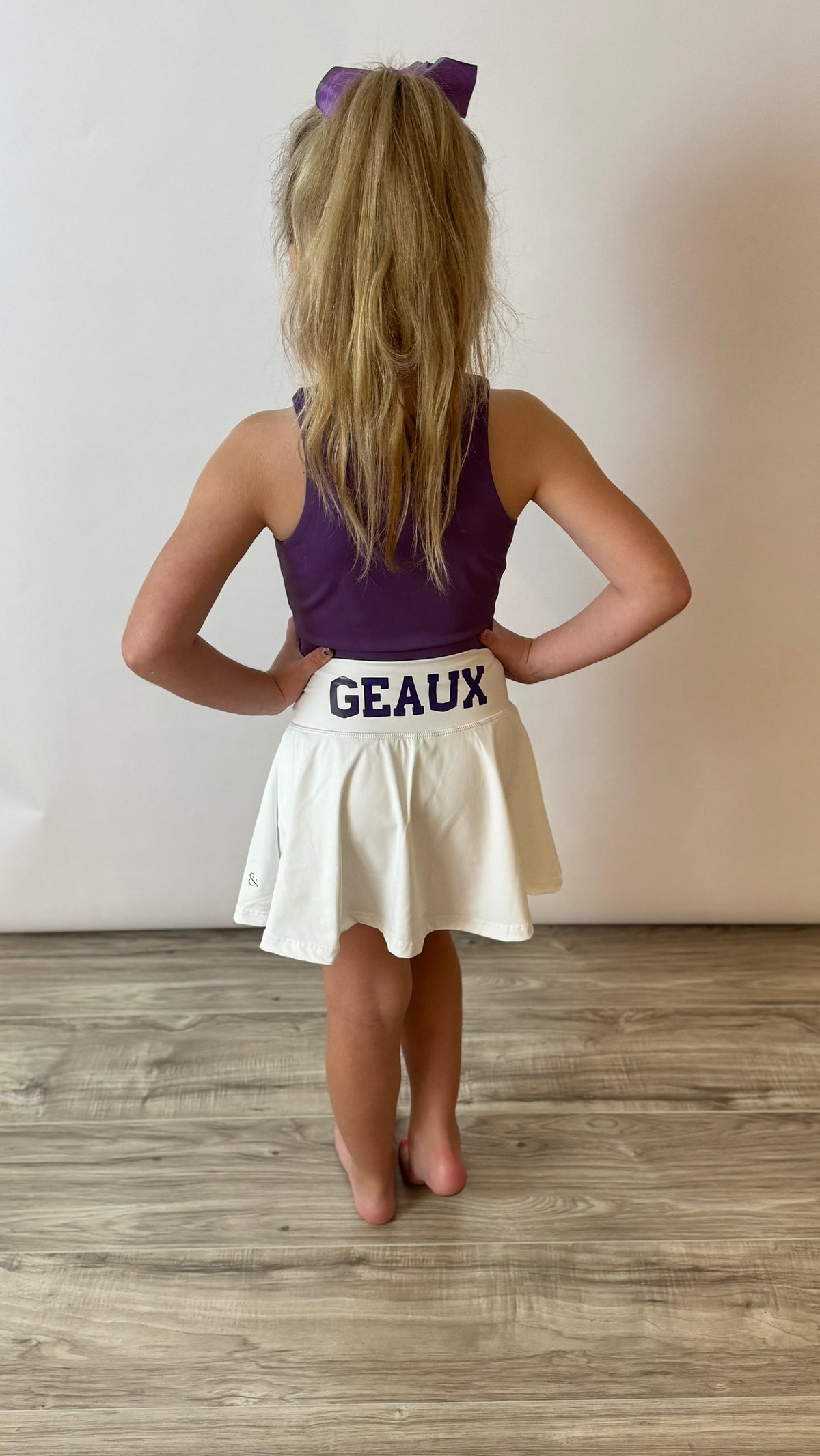 A child wearing a purple and white tennis skirt with 'Geaux' written on the front, and a matching bow. The child has blonde hair and is standing against a white background. This skirt is a game day skirt popular in Louisiana. 