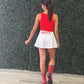 A woman shown from behind wearing a red sleeveless top and white pleated mini skirt with 'HOTTY' printed in red on the back of the skirt, paired with red cowboy boots, standing in front of a green leaf wall.