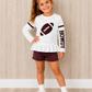 A young girl stands in a room with light wood-paneled walls wearing a white long-sleeve ruffled top with a football graphic and 'HOWDY' text, maroon shorts, white socks, and white sneakers. This is a Gameday shirt. 