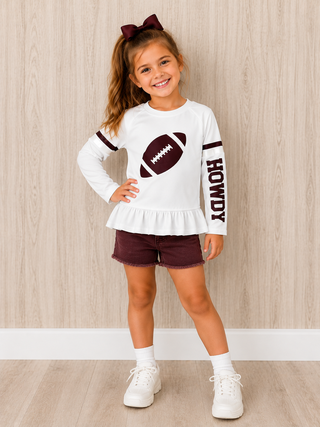 A young girl stands in a room with light wood-paneled walls wearing a white long-sleeve ruffled top with a football graphic and 'HOWDY' text, maroon shorts, white socks, and white sneakers. This is a Gameday shirt. 