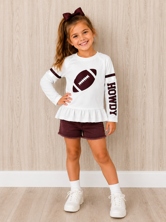 A young girl stands in a room with light wood-paneled walls wearing a white long-sleeve ruffled top with a football graphic and 'HOWDY' text, maroon shorts, white socks, and white sneakers. This is a Gameday shirt. 