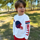 A young boy wearing a white long-sleeve t-shirt with a navy and red football helmet graphic on the chest and the word 'HOTTY' printed down the sleeve, paired with red shorts, standing outdoors in a park. This is a gameday shirt that provides protection from the sun and feels like a t-shirt. 