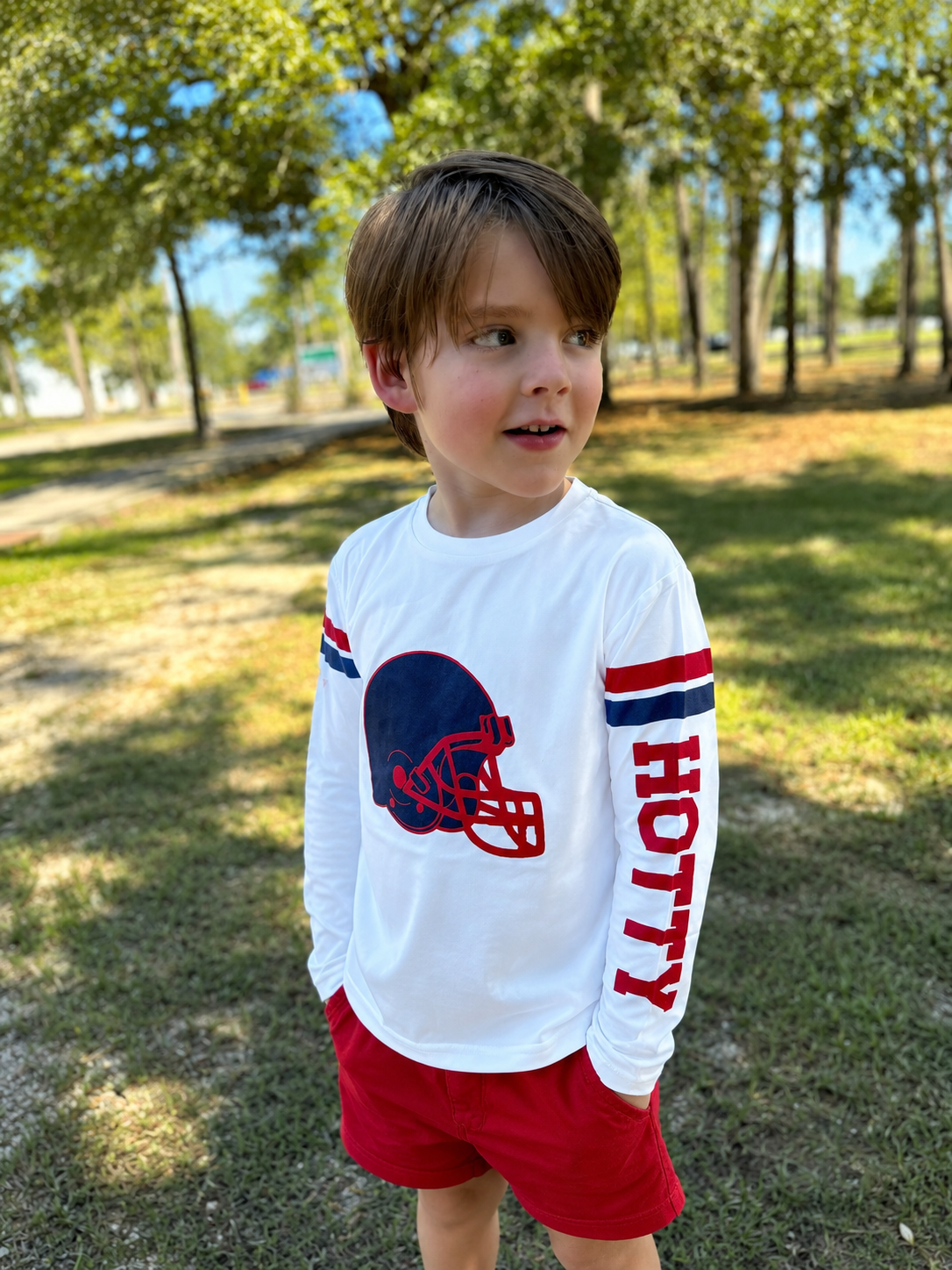 A young boy wearing a white long-sleeve t-shirt with a navy and red football helmet graphic on the chest and the word 'HOTTY' printed down the sleeve, paired with red shorts, standing outdoors in a park. This is a gameday shirt that provides protection from the sun and feels like a t-shirt. 