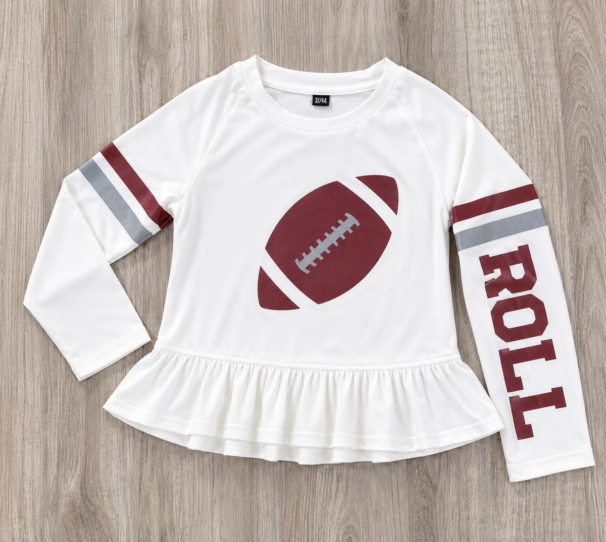 A white long-sleeve t-shirt with a ruffled hem, featuring a crimson football graphic on the chest, crimson and gray stripes on the sleeves, and the word 'ROLL' printed vertically on one sleeve, laid flat on a gray wood background. This is a SPF t-shirt for girls for gameday in Alabama. 