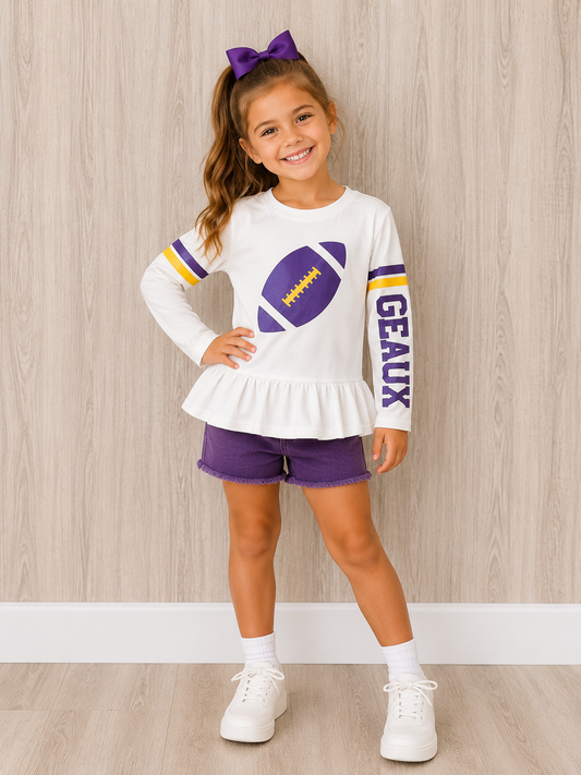 A young girl modeling a white long-sleeve ruffle hem top with purple and yellow football graphics, paired with purple shorts, white socks, and white sneakers. She is standing against a light wood-paneled wall. This a gameday shirt made to worn for protection from the sun while tailgating in Louisiana. 