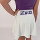 Girl wearing a white skirt with 'GEAUX' text and a purple top on a light pink background