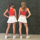Two women standing side by side from the back, wearing matching red tank tops and white pleated skirts with 'HOTTY' printed in red on the back waist. They are standing on a light wood floor in front of a green textured wall.