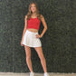 A young woman stands in front of a green artificial hedge wall wearing a red ribbed crop top, a white pleated tennis skirt, and white sneakers. Hotty is written on the back made for gameday. 
