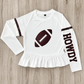 A white long-sleeve athletic SPF top with a ruffled hem, featuring a maroon football graphic with shimmer on the chest, maroon stripes on the sleeves, and 'HOWDY' printed vertically on one sleeve, laid flat on a light wood background. This is a gameday shirt made to be worn in the sun for kids protection. 