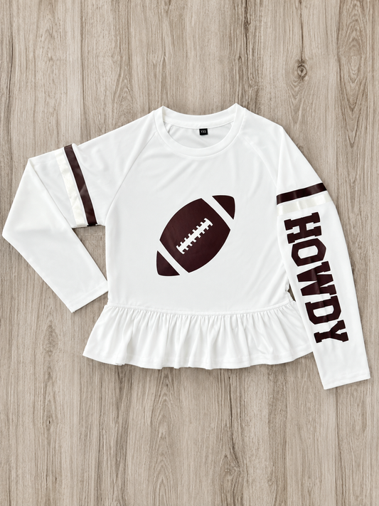 A white long-sleeve athletic SPF top with a ruffled hem, featuring a maroon football graphic with shimmer on the chest, maroon stripes on the sleeves, and 'HOWDY' printed vertically on one sleeve, laid flat on a light wood background. This is a gameday shirt made to be worn in the sun for kids protection. 