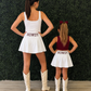 A woman and a child standing with their backs to the camera in matching white mini skirts with 'HOWDY' printed on the back, paired with white cowboy boots. The woman wears a white tank top and the child wears a burgundy tank top, set against a green hedge wall and light wood floor. Howdy is a popular phrase in Texas. 