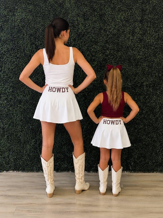 A woman and a child standing with their backs to the camera in matching white mini skirts with 'HOWDY' printed on the back, paired with white cowboy boots. The woman wears a white tank top and the child wears a burgundy tank top, set against a green hedge wall and light wood floor. Howdy is a popular phrase in Texas. 