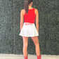 A woman stands with her back to the camera wearing a red sleeveless top, a white pleated mini skirt with 'HOTTY' printed in red on the back, and red cowboy boots, set against a green hedge wall and light wood floor. Mississippi residents know the game day phrase hotty 
