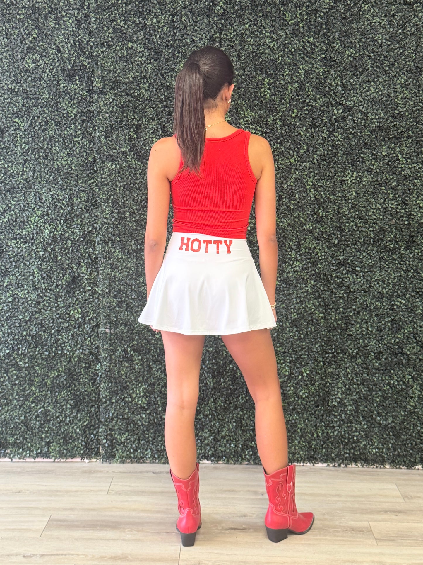 A woman stands with her back to the camera wearing a red sleeveless top, a white pleated mini skirt with 'HOTTY' printed in red on the back, and red cowboy boots, set against a green hedge wall and light wood floor. Mississippi residents know the game day phrase hotty 