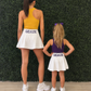 An adult woman and a young girl standing side by side from behind, wearing matching white skater skirts with 'GEAUX' printed in purple on the waistband, along with athletic tops in yellow and purple. They are posed in front of a green artificial hedge wall on a light wood floor. Geaux for Louisiana gameday. 