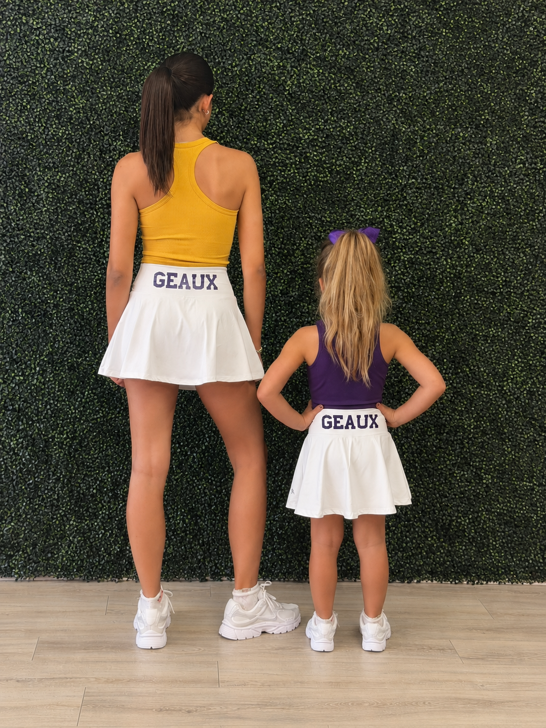 An adult woman and a young girl standing side by side from behind, wearing matching white skater skirts with 'GEAUX' printed in purple on the waistband, along with athletic tops in yellow and purple. They are posed in front of a green artificial hedge wall on a light wood floor. Geaux for Louisiana gameday. 