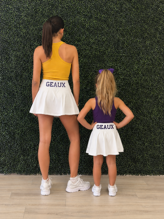 An adult woman and a young girl standing side by side from behind, wearing matching white skater skirts with 'GEAUX' printed in purple on the waistband, along with athletic tops in yellow and purple. They are posed in front of a green artificial hedge wall on a light wood floor. Geaux for Louisiana gameday. 