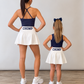 An adult woman and a young girl standing with their backs to the camera in matching navy blue and white cheerleader outfits with 'CHOMP' printed on the back of the skirts, in a studio setting with a light gray wall and wood floor. A gameday matching set for Florida chomp  fans. 