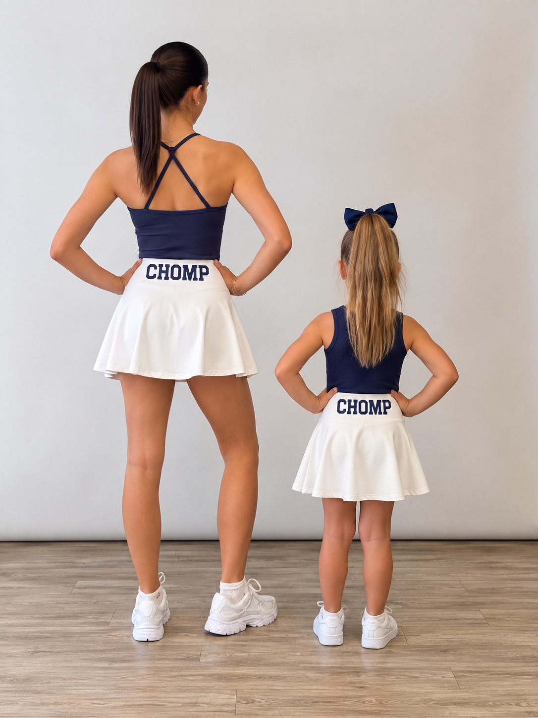 An adult woman and a young girl standing with their backs to the camera in matching navy blue and white cheerleader outfits with 'CHOMP' printed on the back of the skirts, in a studio setting with a light gray wall and wood floor. A gameday matching set for Florida chomp  fans. 