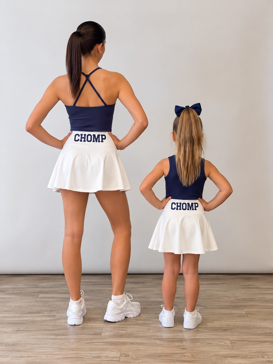 An adult woman and a young girl standing with their backs to the camera in matching navy blue and white cheerleader outfits with 'CHOMP' printed on the back of the skirts, in a studio setting with a light gray wall and wood floor. A gameday matching set for Florida chomp  fans. 