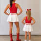 A woman and a young girl standing with their backs to the camera, wearing matching red tank tops, white mini skirts with 'HOTTY' printed in red on the back, and red cowboy boots versus white sneakers, against a plain light gray wall and wood floor. Mississippi Hotty gameday matching skirt sets. 