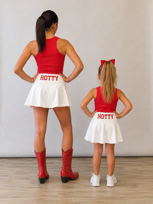 A woman and a young girl standing with their backs to the camera, wearing matching red tank tops, white mini skirts with 'HOTTY' printed in red on the back, and red cowboy boots versus white sneakers, against a plain light gray wall and wood floor. Mississippi Hotty gameday matching skirt sets. 