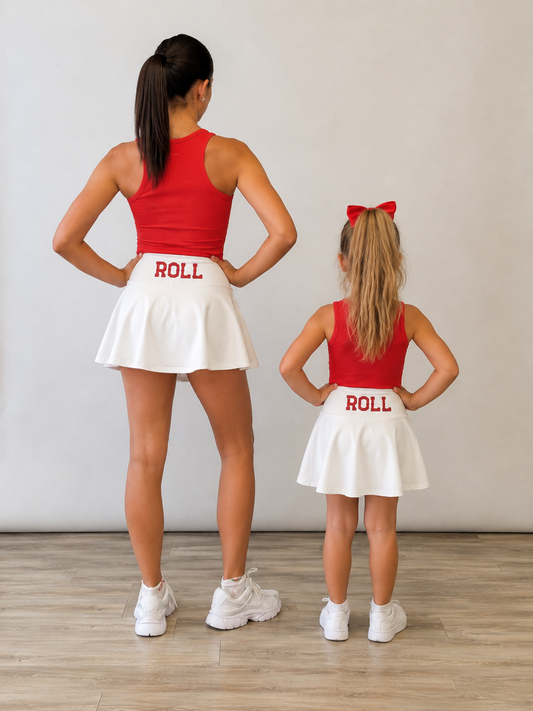 An adult woman and a young girl standing with their backs to the camera, both wearing matching red sleeveless tops and white flared skirts with the word "ROLL" printed in red on the back, posed in a studio with a light gray wall and wood floor. Gameday matching sets. 