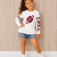 A young girl with a brown hair bow stands against a light wood-paneled wall wearing a white long-sleeve t-shirt with a football graphic and 'ROLL' text, paired with denim shorts, white socks, and white sneakers. The gameday shirt is for fans in Alabama who like football. 