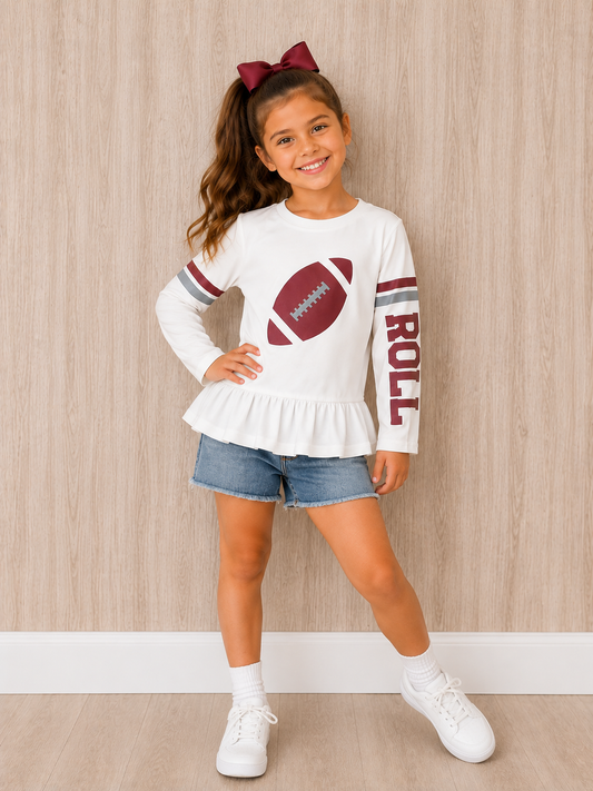 A young girl with a brown hair bow stands against a light wood-paneled wall wearing a white long-sleeve t-shirt with a football graphic and 'ROLL' text, paired with denim shorts, white socks, and white sneakers. The gameday shirt is for fans in Alabama who like football. 