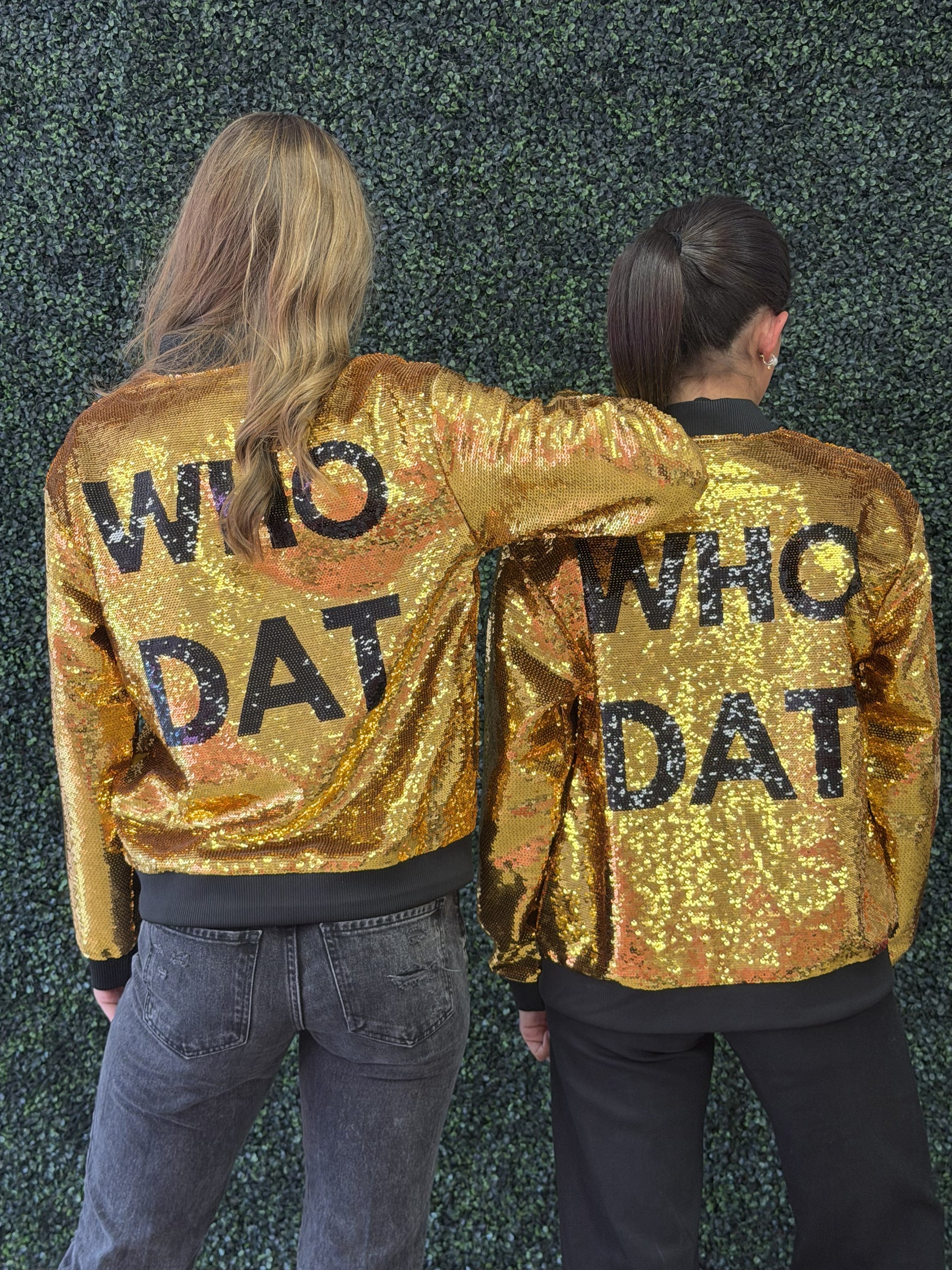 Two women standing with their backs to the camera, wearing matching gold sequined bomber jackets with 'WHO DAT' printed in black across the back, posed against a green textured wall. For New Orleans fans. 