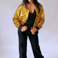 A woman modeling a shiny gold sequined bomber jacket over a black top and black wide-leg pants, standing against a plain light background. The back of the sequin jacket says Who Dat on it. 