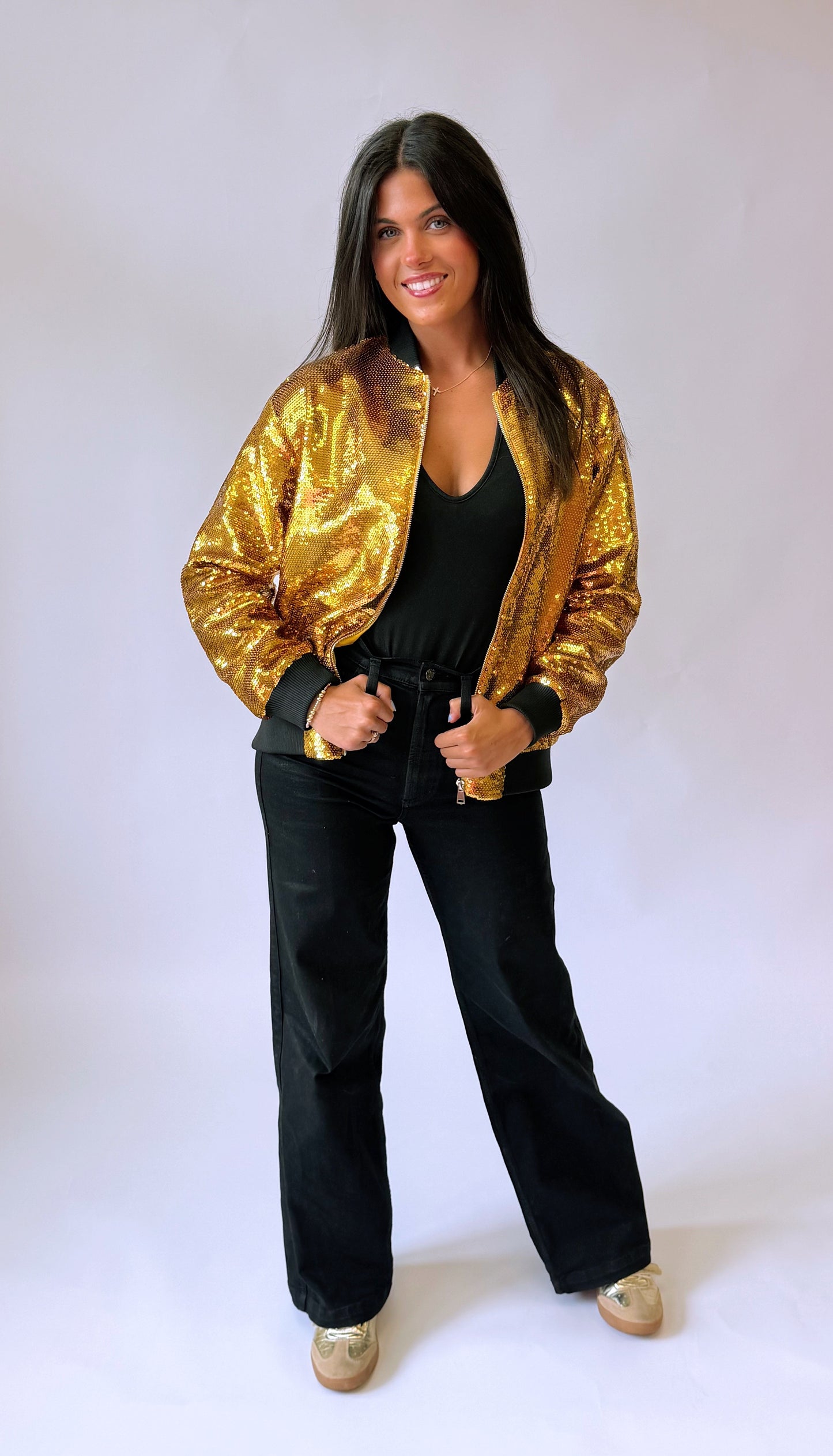 A woman modeling a shiny gold sequined bomber jacket over a black top and black wide-leg pants, standing against a plain light background. The back of the sequin jacket says Who Dat on it. 