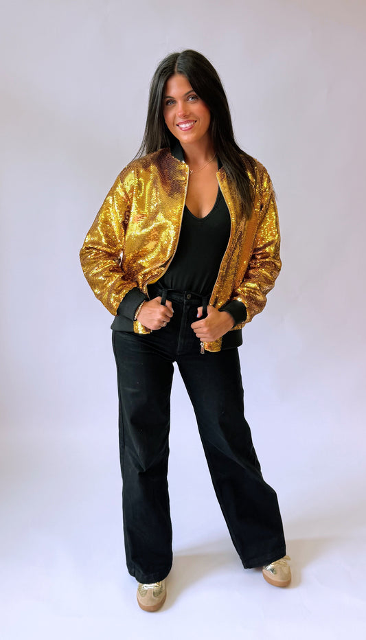 A woman modeling a shiny gold sequined bomber jacket over a black top and black wide-leg pants, standing against a plain light background. The back of the sequin jacket says Who Dat on it. 
