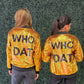 Two women shown from the back wearing matching gold sequined bomber jackets with 'WHO DAT' printed in black across the back, standing against a green hedge wall. 