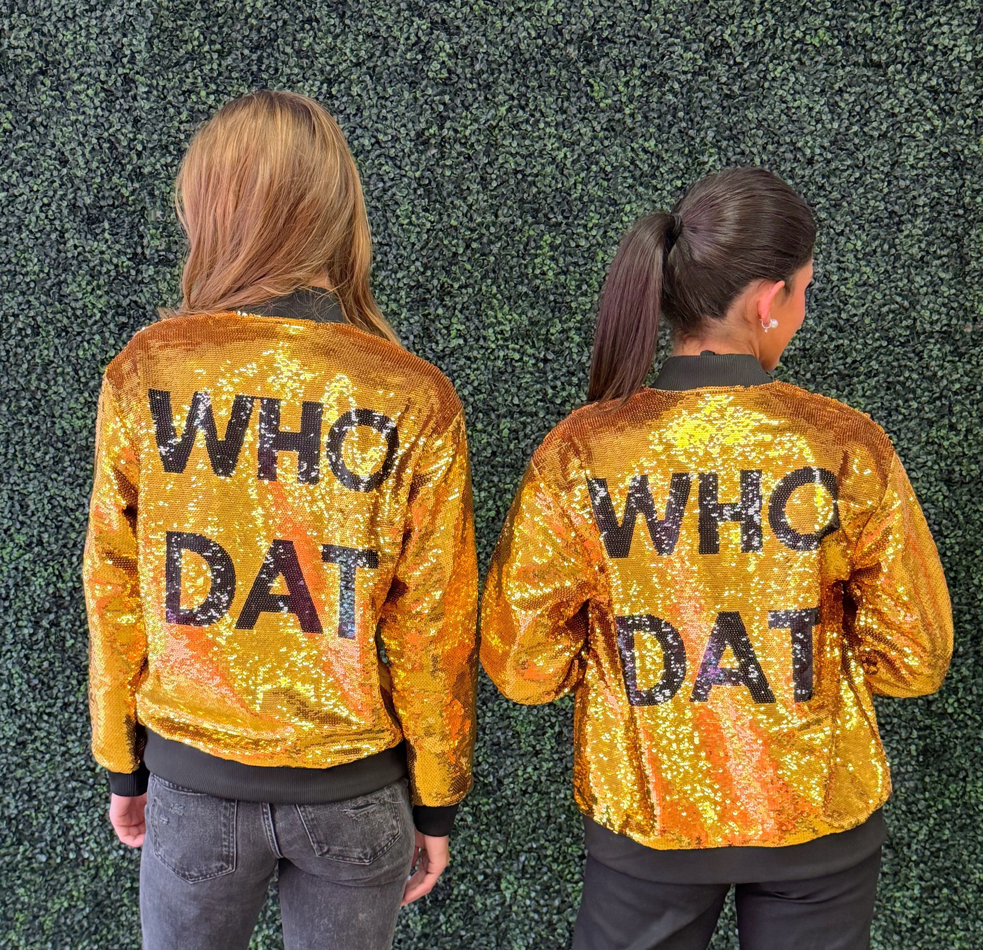 Two women shown from the back wearing matching gold sequined bomber jackets with 'WHO DAT' printed in black across the back, standing against a green hedge wall. 