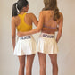 Two women standing side by side from behind wearing matching white game day athletic tennis skirts pleated skirts with 'GEAUX' printed in purple on the back, along with purple and golds sports bras, against a plain light beige studio background.