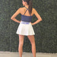 A woman stands with her back to the camera wearing a navy blue halter crop top and a white tennis skirt with 'CHOMP' printed in blue on the waistband. She is posing in front of a green artificial hedge wall and light wood flooring. 