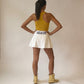 A woman stands with her back to the camera wearing a yellow racerback tank top, a white pleated athletic tennis skirt with 'GEAUX' printed in purple  on the waistband, and gold boots, set against a plain beige studio backdrop.