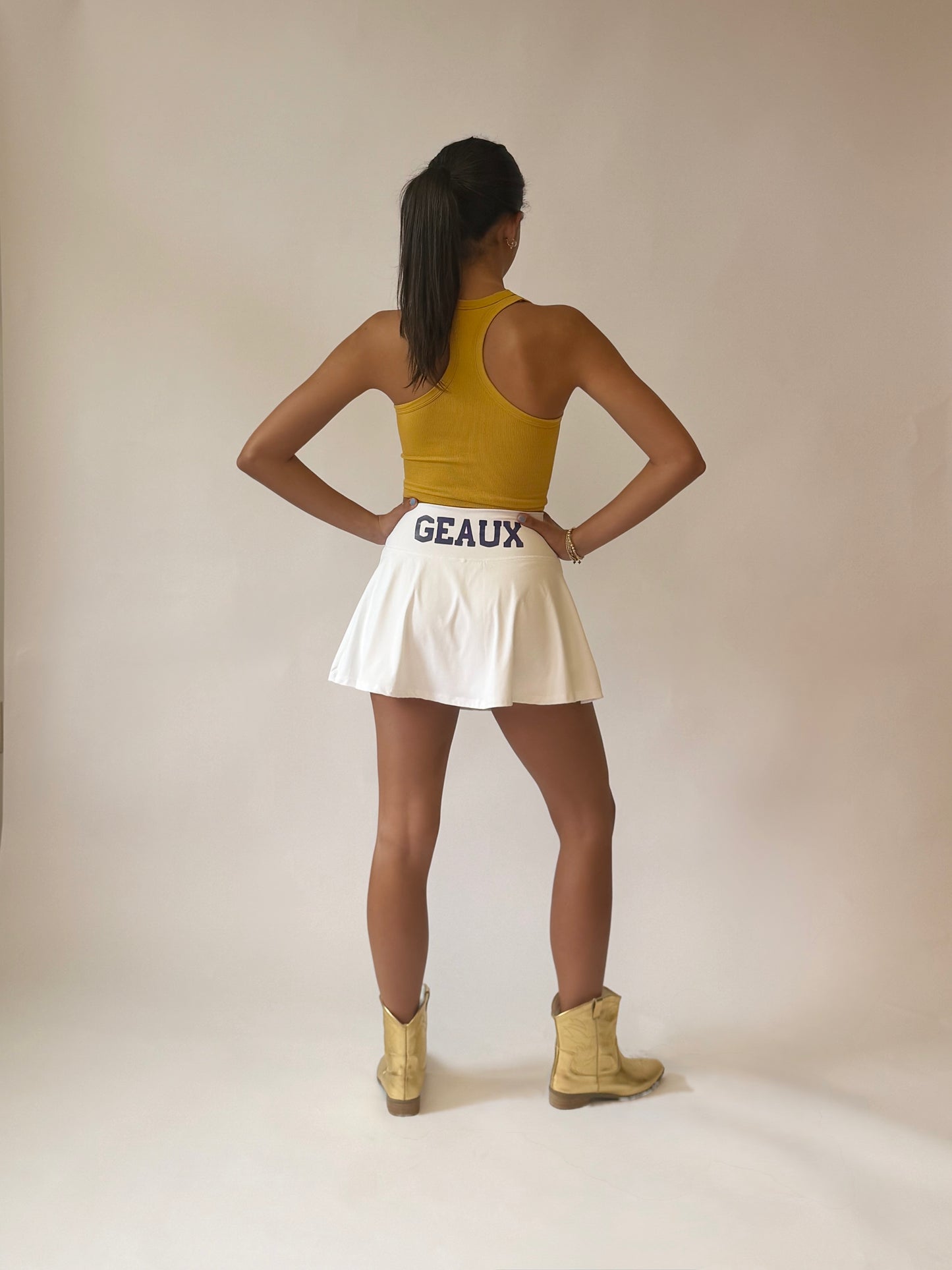 A woman stands with her back to the camera wearing a yellow racerback tank top, a white pleated athletic tennis skirt with 'GEAUX' printed in purple  on the waistband, and gold boots, set against a plain beige studio backdrop.