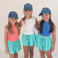 Three young girls wearing matching blue caps and turquoise shorts on a plain background. Tween kids trucker hats with fearless written on them. 