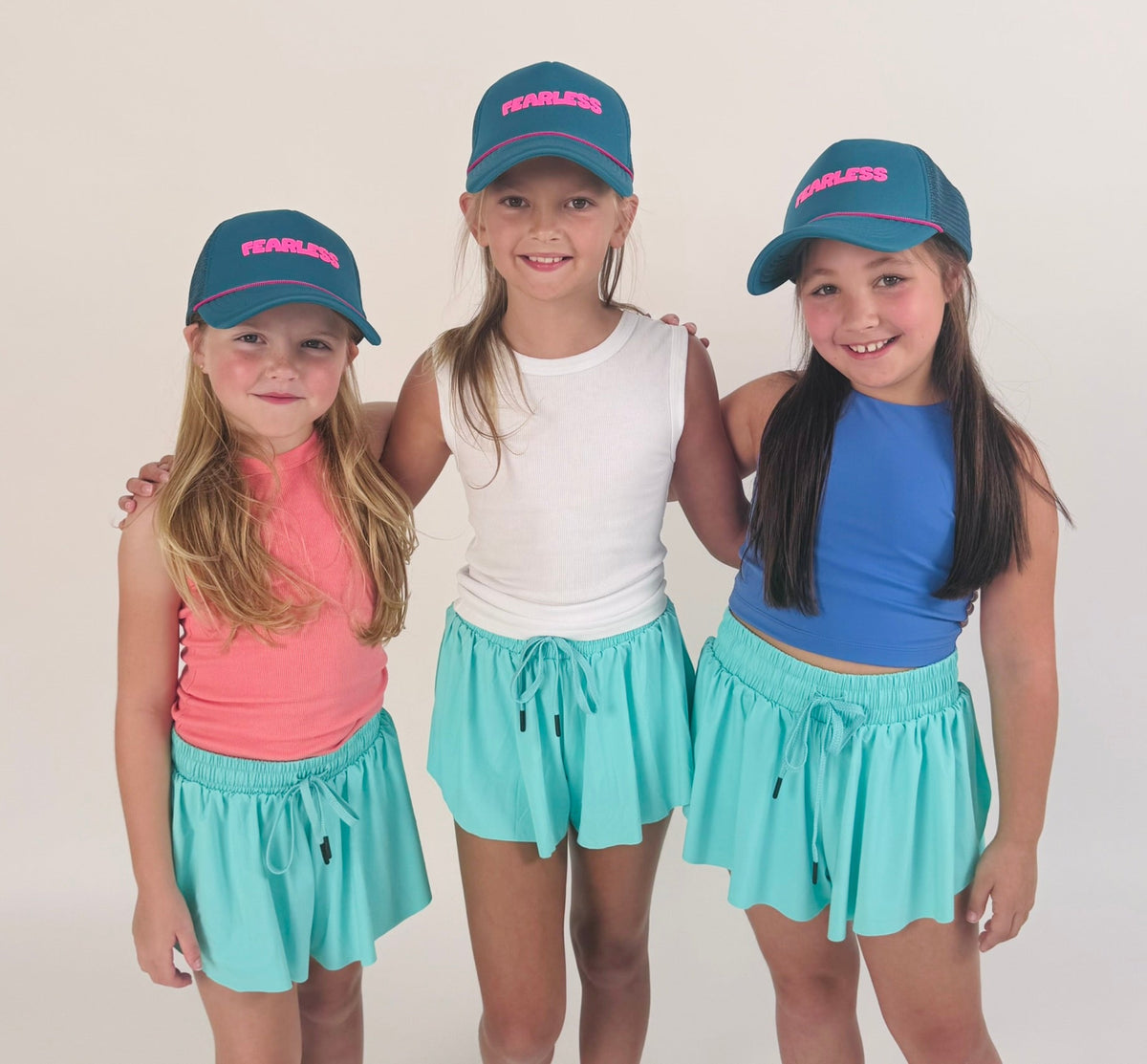 Three young girls wearing matching blue caps and turquoise shorts on a plain background. Tween kids trucker hats with fearless written on them. 